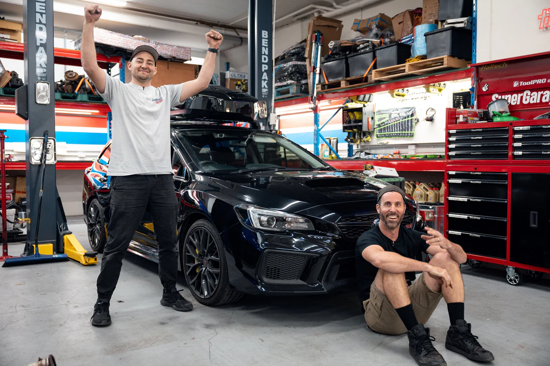 Two automotive mechanics celebrating successful performance clutch installation on dark blue sports car in professional workshop garage with Xtreme Performance branding